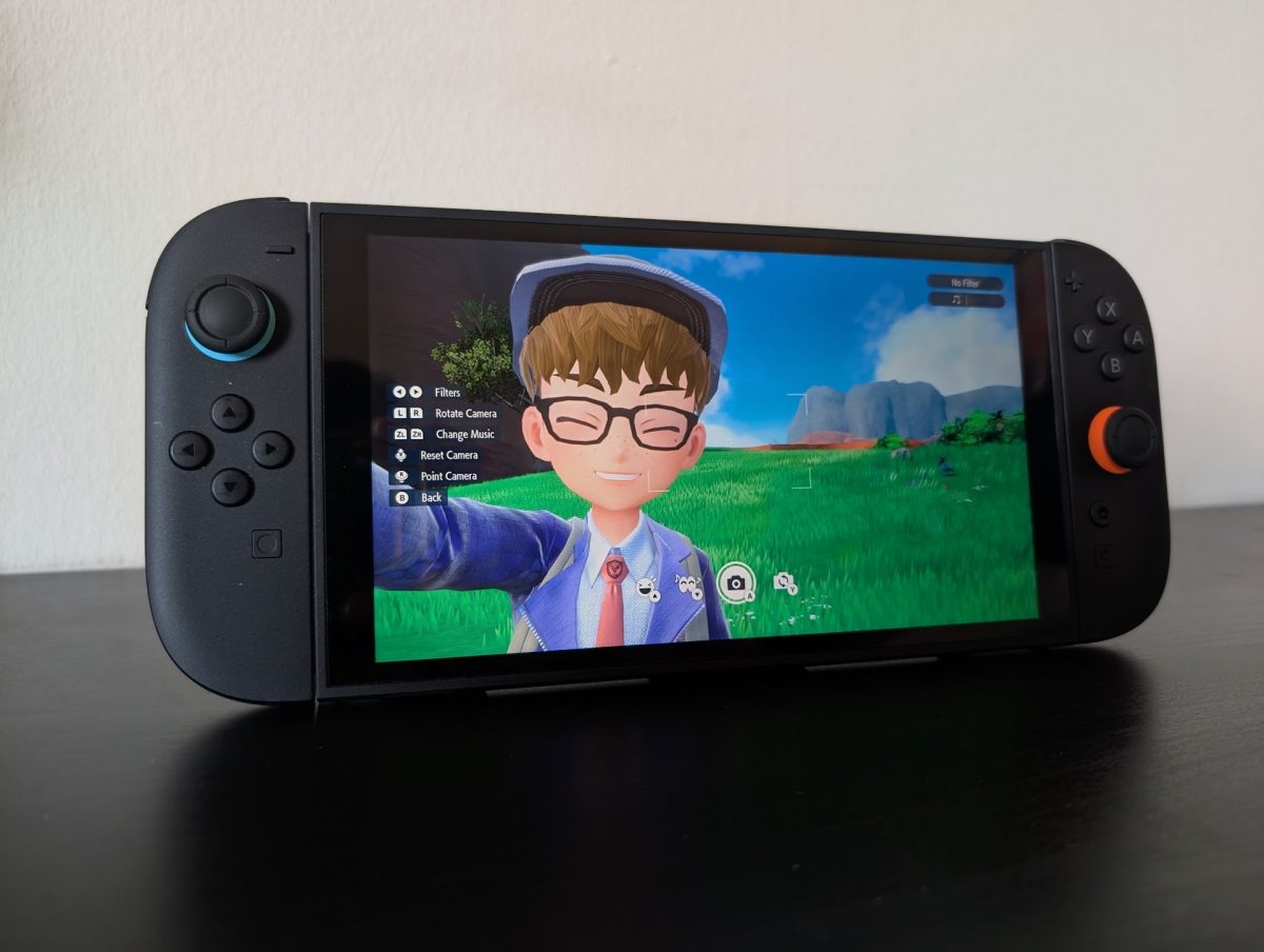 Un entraîneur prend un selfie dans Pokémon Écarlate et Violet, tournant sur une Switch 2 sur une table noire contre un mur blanc.